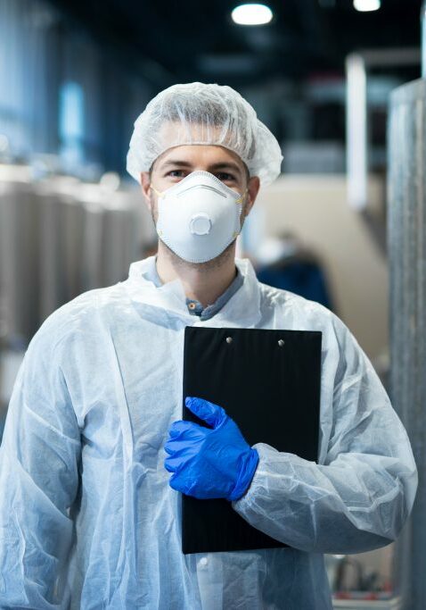 Technologist with protective mask and hairnet standing at factory production line. Food or beverage processing factory.